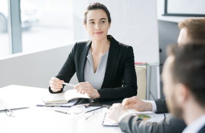 Portrait of confident young businesswoman smiling while listening to colleagues sitting at meeting table in conference room during briefing, copy space